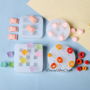 Puede incluir: Una colección de moldes de silicona y alimentos en miniatura. Los moldes blancos contienen caramelos, cupcakes y formas de cereales. Los alimentos en miniatura son de varios colores, incluyendo rosa, naranja y morado. El texto "Sweet" es visible en los envoltorios de caramelos.