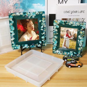 Pode incluir: Duas molduras quadradas turquesa decoradas com conchas, cada uma com uma foto de uma pessoa. Uma bandeja de plástico transparente e dois carros de corrida de brinquedo estão em frente às molduras. As molduras estão em suportes de metal preto.