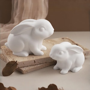 Könnte beinhalten: Zwei weiße Keramik-Hasenfiguren. Einer ist größer und sitzt auf einem strukturierten, rechteckigen Stein. Der andere, kleinere Hase befindet sich daneben. Ein getrocknetes Blatt liegt in der Nähe und fügt der Szene ein natürliches Element hinzu.