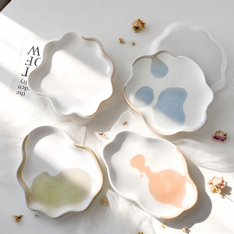 May include: Four white ceramic dishes with irregular, wavy edges. One dish has blue abstract paint, another has peach, and a third has green. The fourth dish is plain white. Dried flowers are scattered around the dishes.