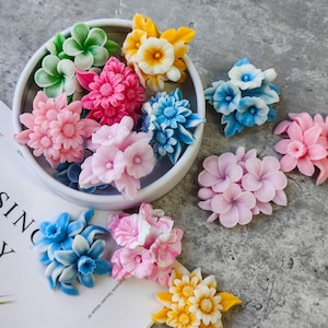 Peut inclure: Un assortiment de savons en forme de fleurs colorées, dans des tons de rose, bleu, jaune et vert. Les savons sont disposés dans un bol blanc et dispersés sur une surface blanche, présentant divers motifs floraux.