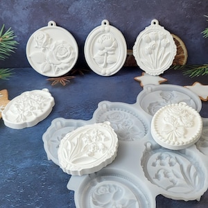 May include: A collection of white silicone molds and finished white plaster ornaments featuring various floral designs including roses, tulips, and lilies. The molds are arranged with several completed circular and oval relief pieces.