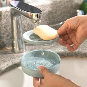 May include: A light beige bar of soap rests on a light blue soap dish, with water dripping into a matching bowl below. The soap dish and bowl are made of a marbled material. A chrome tap is in the background.