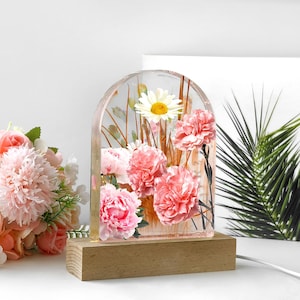 Op de afbeelding: Decoratieve bloemenharslamp met roze anjers, een witte madelief en gedroogde grassen, ingekapseld in een helder, boogvormig harsblok. De lamp staat op een rechthoekige houten basis.