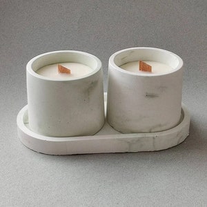 May include: Two white candles in cylindrical concrete holders with wooden wicks, set on a matching oval tray. The holders and tray have a marbled effect. The candles are unscented and designed for home decor.