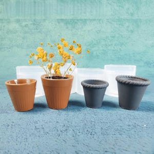 May include: An assortment of small terracotta and gray ceramic flower pots. One pot contains yellow flowers. The pots vary in size and design, with ribbed and rope-edged details. Clear plastic molds are visible in the background.