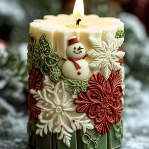 May include: A lit, ivory-colored pillar candle with a snowman and snowflake design. The candle features a snowman with a red scarf and hat, surrounded by green, red, and white snowflake and floral accents. The base is green.
