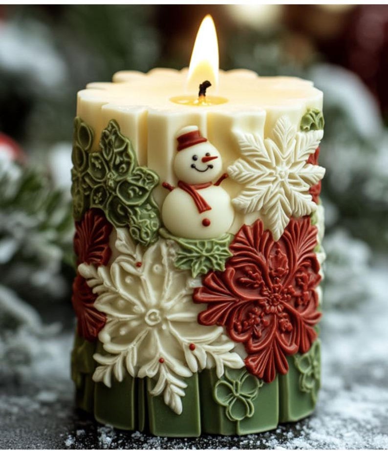 May include: A lit, ivory-colored pillar candle with a snowman and snowflake design. The candle features a snowman with a red scarf and hat, surrounded by green, red, and white snowflake and floral accents. The base is green.