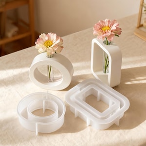 May include: White ceramic bud vases in oval and square shapes, each holding a single flower. Two additional white geometric molds are also present. The flowers are pink with yellow centers. The vases are on a cream-colored surface.