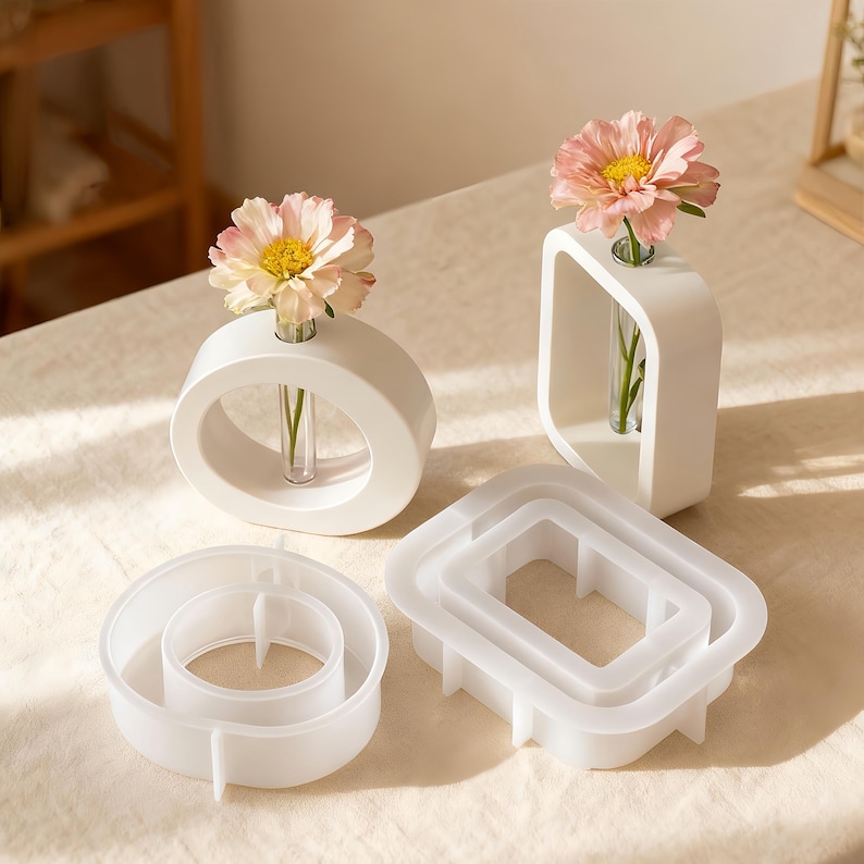 May include: White ceramic bud vases in oval and square shapes, each holding a single flower. Two additional white geometric molds are also present. The flowers are pink with yellow centers. The vases are on a cream-colored surface.