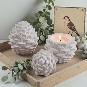 Peut inclure: Ensemble de décorations blanches en forme de pommes de pin, dont un bougeoir avec une bougie allumée, disposées sur un plateau en bois. Une illustration d'oiseau encadrée et de la verdure artificielle complètent la scène.