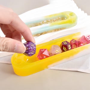 Op de afbeelding: Een verzameling kleurrijke polyhedrale dobbelstenen in een gele en witte harsbak. De dobbelstenen zijn in verschillende kleuren, waaronder paars, rood en roze, met gouden cijfers. Op de achtergrond staat een andere bak met een blauw en geel ontwerp.