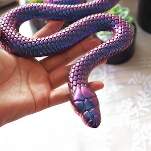 Könnte beinhalten: Eine flexible, bewegliche Schlangenfigur mit einer metallisch schimmernden Oberfläche, die zwischen tiefen Violett- und Blautönen wechselt. Die Oberfläche ist mit einem detaillierten, erhabenen Schuppenmuster versehen und der Kopf zeigt definierte, segmentierte Platten.