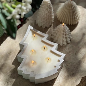 May include: A white, Christmas tree-shaped candle with six lit wicks. Three smaller, white tree-shaped candles are in the background. The candles are on a light brown, textured surface. The candles are a festive, decorative item.