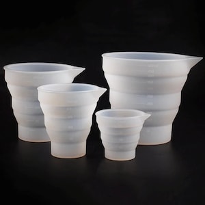 May include: Four white, collapsible measuring cups of different sizes are displayed against a black backdrop. Each cup features a pouring spout and milliliter measurement markings. The cups are made of a flexible material.