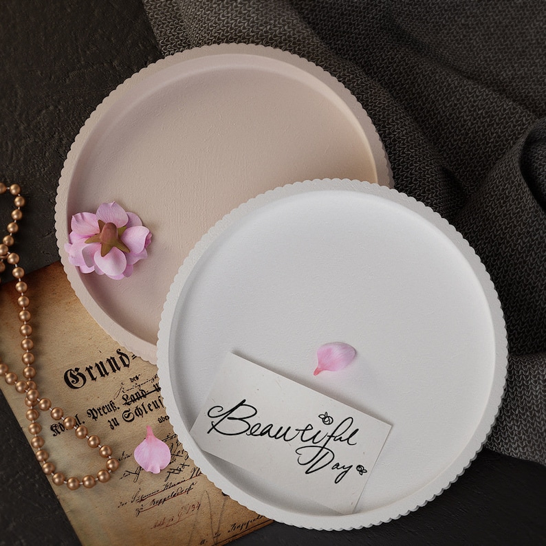 Pu&ograve; includere: Due vassoi decorativi rotondi, uno rosa chiaro e uno bianco, con bordi smerlati. Un fiore rosa e una carta con le parole "Beautiful Day" sono sui vassoi. Una collana d'oro e un pezzo di carta con testo sono anche nell'immagine.
