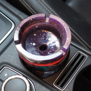 Puede incluir: Un cenicero redondo, morado y plateado con un dise&ntilde;o de galaxia, con estrellas y purpurina. El cenicero de resina tiene forma circular con un agujero central. Est&aacute; colocado en la consola de un coche.