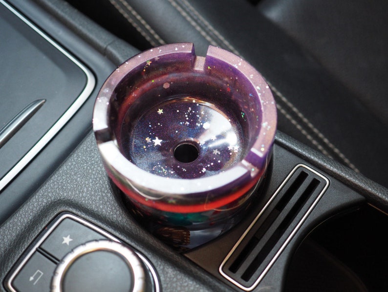 Puede incluir: Un cenicero redondo, morado y plateado con un dise&ntilde;o de galaxia, con estrellas y purpurina. El cenicero de resina tiene forma circular con un agujero central. Est&aacute; colocado en la consola de un coche.