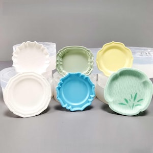 May include: A collection of small, decorative plates in various pastel colors. The plates have unique shapes, including scalloped edges and floral designs. Colors include white, green, yellow, and blue. These plates are likely for serving small portions or decorative use.