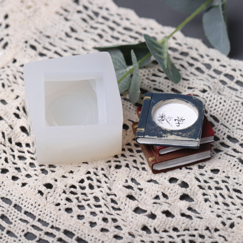 May include: A white silicone mold with a square shape and a circular indentation. Stacked miniature books in blue, red, and brown are also visible. The top book has text inside a circular opening.