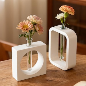 May include: Two white, geometric vases with clear glass tubes holding flowers. One vase is square with a circular cutout, the other is rectangular with a curved top. The flowers are peach and white.