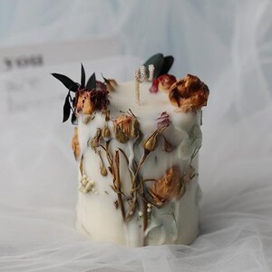 May include: A white, square candle decorated with dried flowers and foliage. The candle features a wick and is adorned with various dried flowers, buds, and leaves in shades of brown, red, and green. The candle is set against a soft, white background.