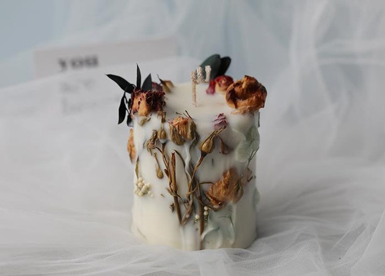 May include: A white, square candle decorated with dried flowers and foliage. The candle features a wick and is adorned with various dried flowers, buds, and leaves in shades of brown, red, and green. The candle is set against a soft, white background.
