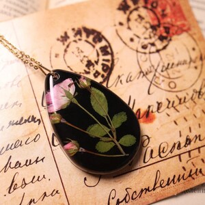 May include: A gold chain necklace with a teardrop-shaped pendant. The black pendant features a preserved pink rose and green leaves. The necklace is displayed on a vintage paper background with handwritten text and stamps.