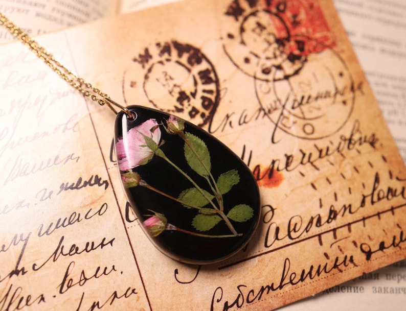 May include: A gold chain necklace with a teardrop-shaped pendant. The black pendant features a preserved pink rose and green leaves. The necklace is displayed on a vintage paper background with handwritten text and stamps.
