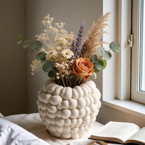 May include: A cream-colored ceramic vase with a textured, bubble-like surface holds a dried floral arrangement. The bouquet includes a single orange rose, white daisies, purple lavender, feathery pampas grass, and green eucalyptus leaves.