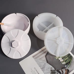 Puede incluir: Ceniceros blancos con tapas. Un cenicero tiene un cigarrillo. Las tapas tienen un diseño geométrico. Los ceniceros son redondos y tienen un interior dividido. Los ceniceros están sobre una superficie gris.