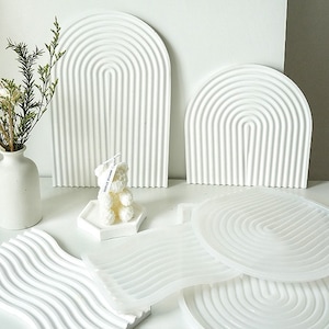 May include: A selection of white decorative pieces, including arched wall art, wave-patterned trays, and a bear-shaped candle. A white vase holds dried flowers. The items are arranged on a white surface, creating a minimalist aesthetic.