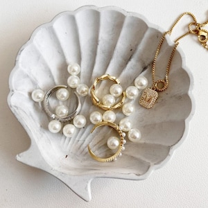 May include: Jewellery displayed in a white seashell-shaped dish. The items include gold and silver rings, a gold necklace with a pendant, and scattered faux pearls. The rings have pearl and crystal accents.