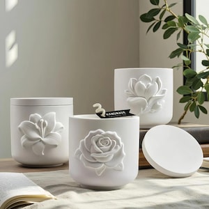 Peut inclure: Trois récipients à bougie cylindriques en céramique blanche avec des motifs floraux en relief. Un récipient présente un lys, un autre une rose et le troisième un magnolia. Une étiquette noire avec le texte "HANDMADE" est fixée à la bougie rose.