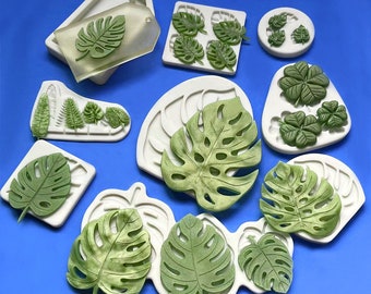Puede incluir: Una variedad de moldes de silicona blancos y decoraciones en forma de hojas verdes. Los moldes presentan diseños detallados de hojas, incluyendo hojas de monstera y helecho. Las decoraciones son de varios tonos de verde, con intrincados detalles de vetas.