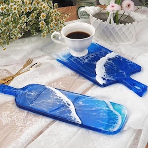 May include: Two blue resin serving boards with white wave designs. One board has a handle and a cup of coffee on it. The other board is a similar shape but without a handle. The boards are on a white lace tablecloth.