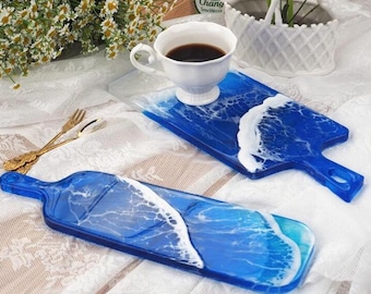 May include: Two blue resin serving boards with white wave designs. One board has a handle and a cup of coffee on it. The other board is a similar shape but without a handle. The boards are on a white lace tablecloth.