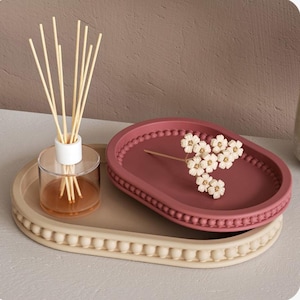 May include: An oval beige tray with a beaded edge holds a smaller maroon tray with a beaded edge and a sprig of white flowers. A clear glass diffuser with reeds sits on the beige tray.