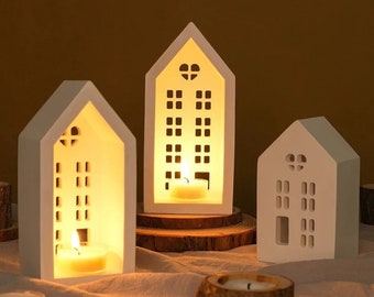 Puede incluir: Tres candelabros blancos de cerámica con forma de casa, con ventanas recortadas y detalles en forma de corazón. Cada casa contiene una vela de té encendida, que emite un brillo cálido. Los candelabros están colocados sobre una superficie neutra.