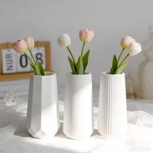 Könnte beinhalten: Ein Set aus drei Keramikbechern mit gesprenkelter, matter Oberfläche. Die Becher sind in Creme-, Hellgrau- und Dunkelgrautönen gehalten. Jeder Becher hat ein schlichtes, minimalistisches Design, ideal für Kaffee oder Tee.