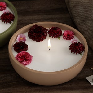 Puede incluir: Una vela encendida en un cuenco de cerámica beige, rodeada de flores flotantes de color rojo oscuro y rosa. La vela tiene una superficie de cera blanca y una sola mecha con una pequeña llama. Otra vela similar en un cuenco verde es visible.