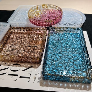 May include: A collection of decorative resin trays in various shapes and colors. The trays include a round pink tray with gold flakes, a square copper tray, and a rectangular blue tray. The trays have a textured, faceted design.