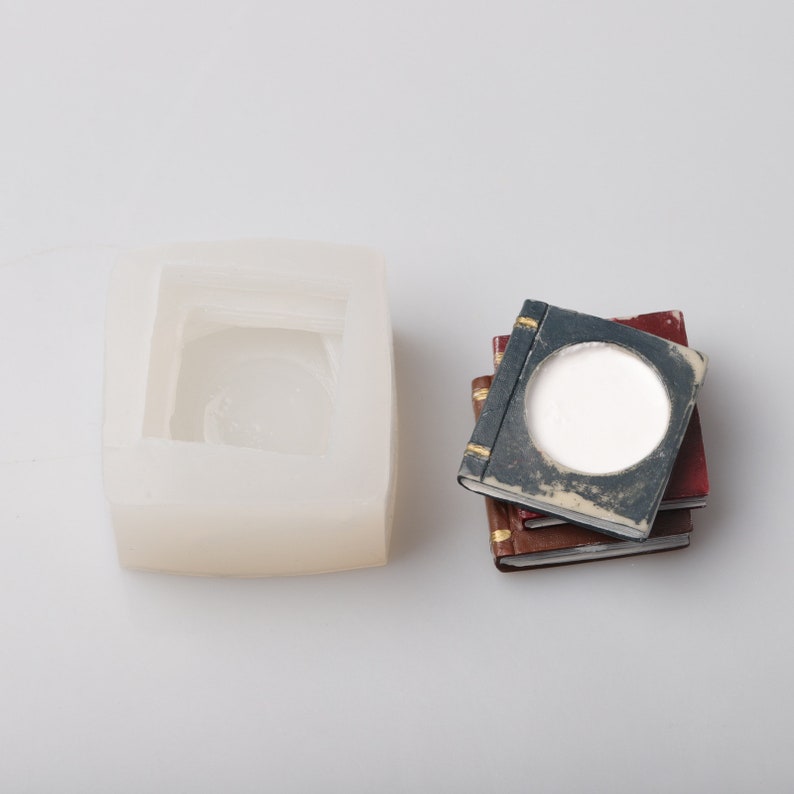 May include: A clear silicone mold for crafting, next to a stack of miniature books. The books have aged, textured covers in shades of blue, red, and brown, with gold accents. One book has a circular opening filled with white material.