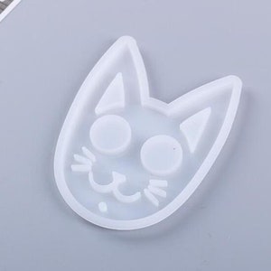 May include: A clear, cat-shaped mold made of flexible silicone. The mold features a cat face with large round eyes, triangular ears, and a smiling mouth with whiskers. The mold is designed for crafting and creating cat-themed items.