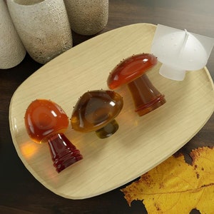 May include: A wooden serving tray holds three glass mushroom-shaped objects in shades of amber and brown, alongside a white mushroom-shaped object. The mushrooms have textured caps and stems. A yellow leaf rests nearby.