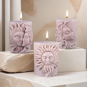 May include: Three lit, lavender-colored pillar candles. Each candle features a carved design: a sun, a sun and moon embracing, and a moon with a face. The candles are on a white surface and a stone. The text "DaisyNoteCraft" is visible.