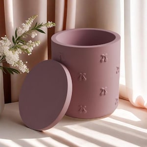 May include: A cylindrical, mauve-colored container with a matching lid. The container features a series of embossed cherry designs. The lid is circular and rests beside the container. The background includes white flowers and a soft, neutral-toned curtain.