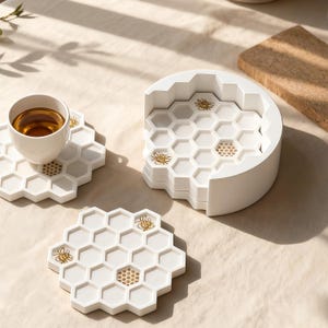 May include: White ceramic honeycomb-shaped coasters with gold bee accents. A stack of coasters is next to a single coaster with a teacup on top. The coasters are designed to resemble a honeycomb.