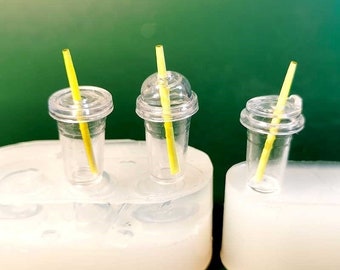 May include: Three miniature clear plastic cups with yellow straws. The cups have clear lids and are set in a white mould. The background is a solid green colour. The cups are small, likely for crafting or dollhouse use.