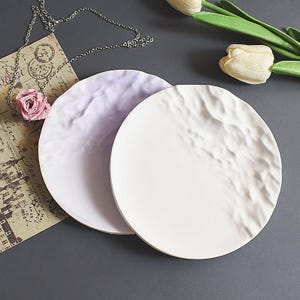 May include: Two round, textured ceramic plates, one white and one pale lavender. A silver necklace with a pink rose pendant and a vintage postcard are in the background. White tulips are also visible.